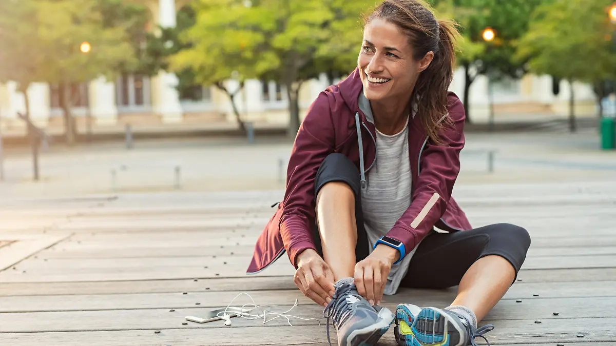 Mature fitness woman tie shoelaces on road. Cheerful runner sitting on floor on city streets with mobile and earphones wearing sport shoes. Active latin woman tying shoe lace before running.