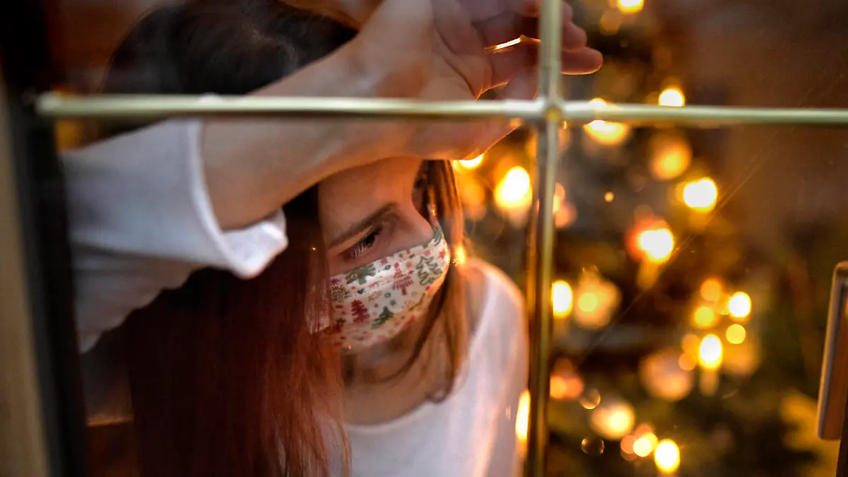 21.11.2020, Modelreleased. Symbolbild, Eine Frau mit Mund-Nasenschutz-Maske schaut sehnsüchtig aus dem Wohnzimmerfenster, innen brennen die kerzen des Weihnachstbaumes, sie ist alleine. Angst vor Einsamkeit wegen Corona am Heiligen Abend. Kontaktverbote und Ausgangsbeschränkungen machen der Bevölkerung das Leben schwer, die gewohnten Treffen unter den Familien könnte es dieses Jahr wegen der Pandemiebeschränkungen nicht geben. Depressionen können die Folge sein. 21.11.2020, Weihnachten in Einsamkeit wegen Corona 21.11.2020, Weihnachten in Einsamkeit wegen Corona *** 21 11 2020, Modelreleased symbol image, A woman with mouth Nose protection Mask looks longingly out of the living room window, inside the candles of the Christmas tree