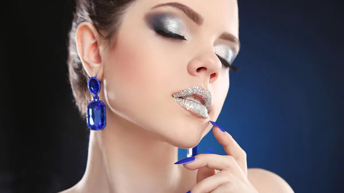Lips. Sparkle Makeup. Closeup of beauty fashion girl with silver lipstick and glitter eyeshadow, wears in blue earrings jewelry over dark studio background.