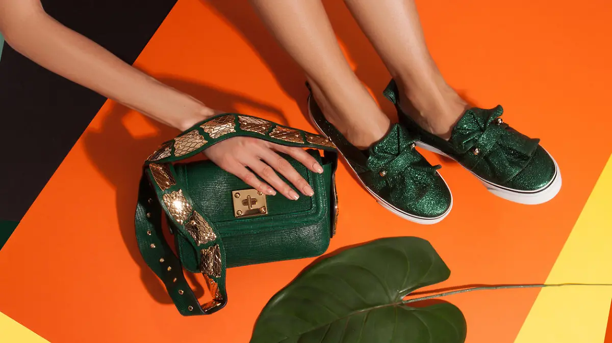 Young woman feet in green snake leathure flat slip-on shoes holding green textured leather metal decorated bag over colored orange yellow and black paper backgroung near palm leaf. Trendy layout