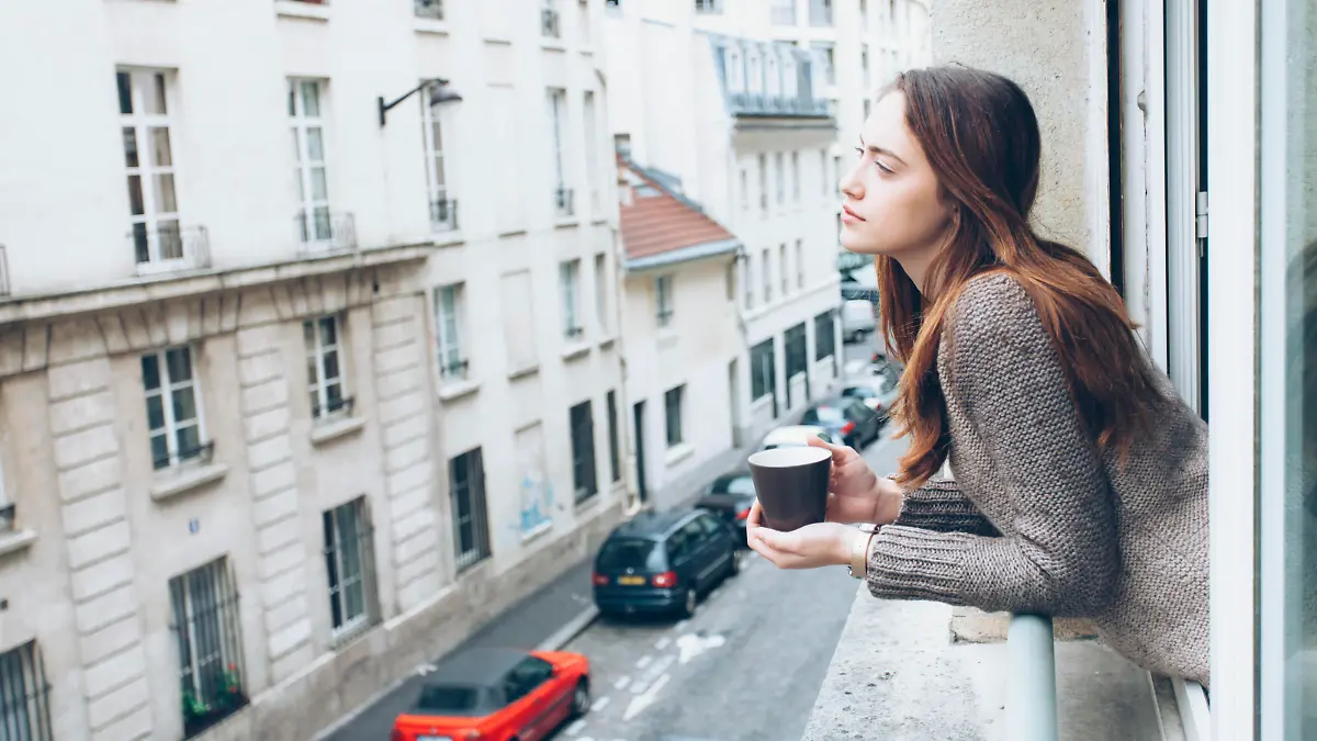 Young woman leaning on window and drinking coffee, looking away. Street view, car and building exterior on background.