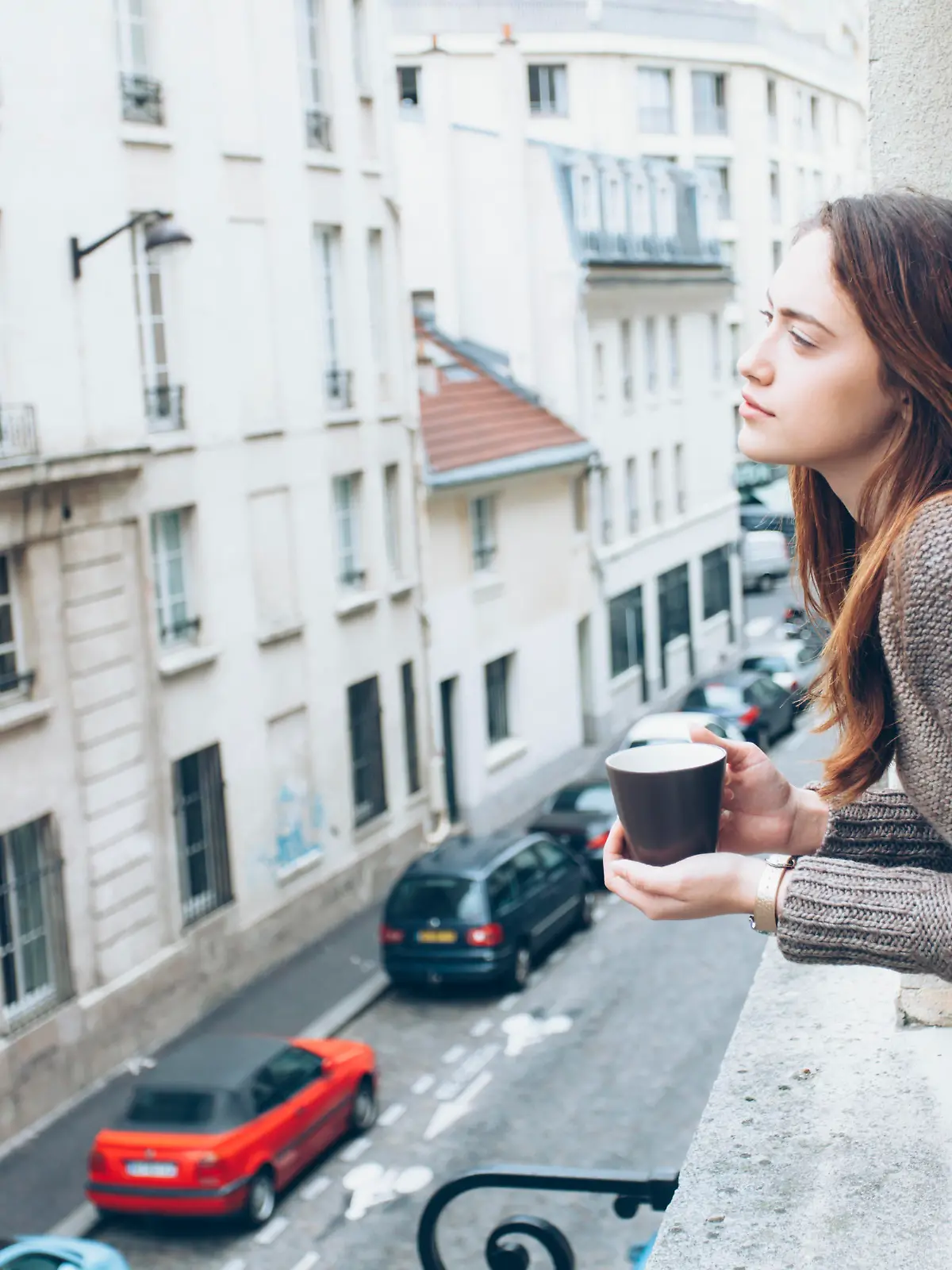Young woman leaning on window and drinking coffee, looking away. Street view, car and building exterior on background.