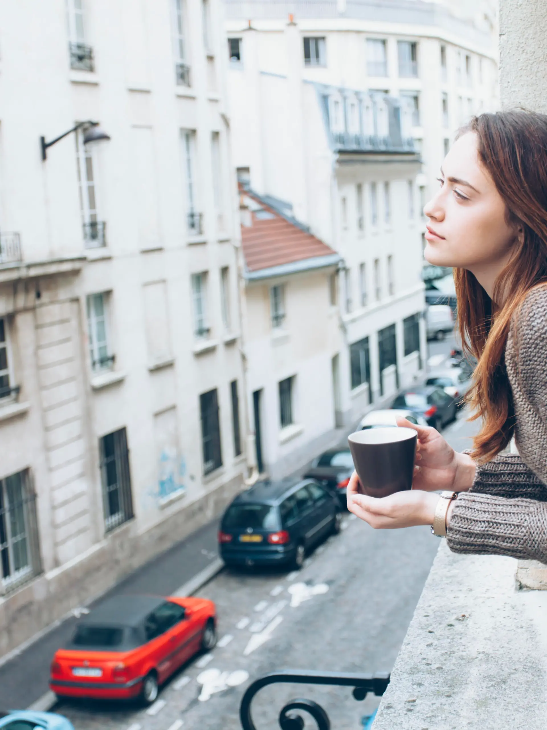 Young woman leaning on window and drinking coffee, looking away. Street view, car and building exterior on background.