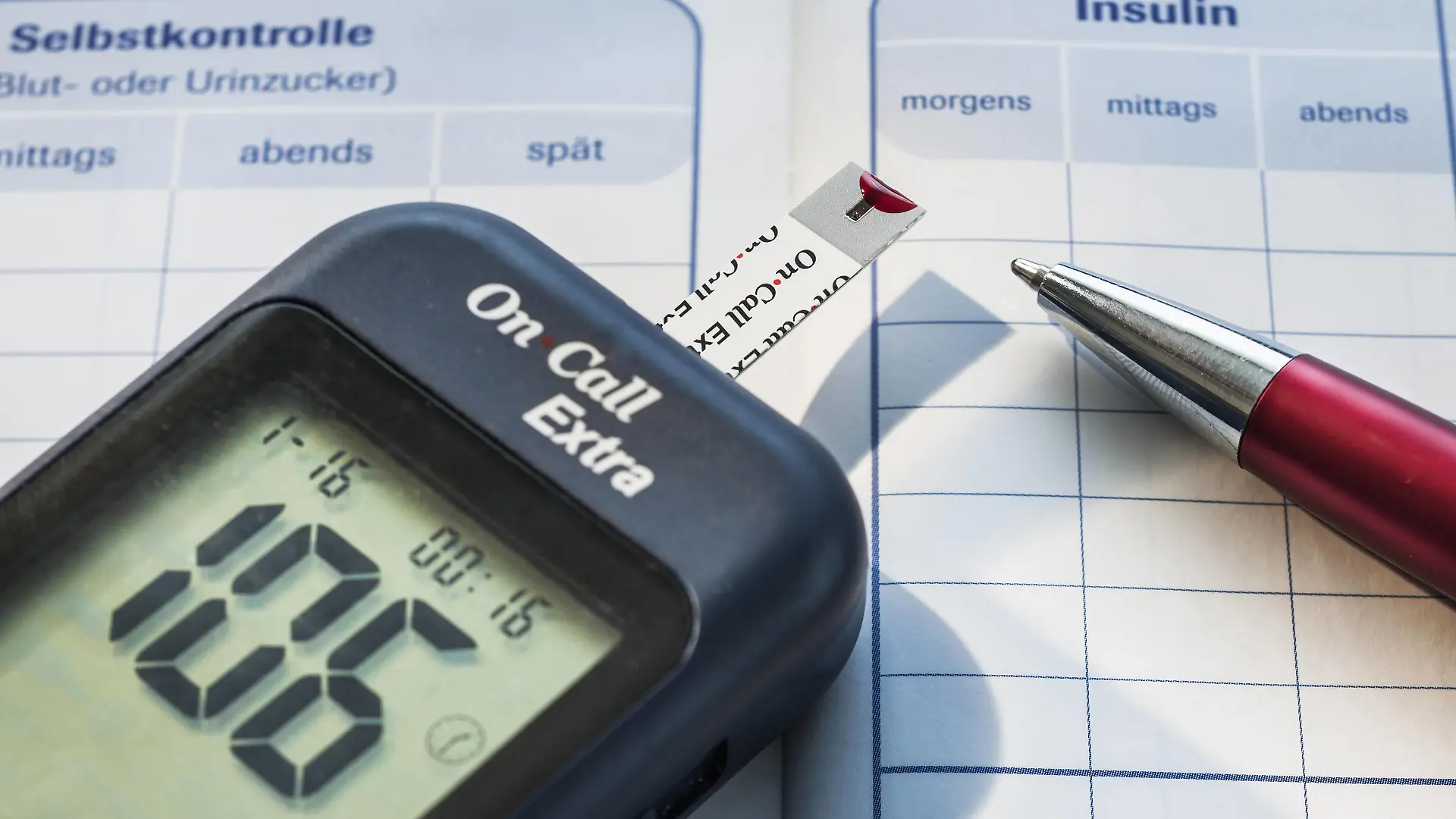  Symbolbild Diabetes, Messgerät zur Blutzuckerbestimmung mit einem Bluttropfen auf dem Messstreifen liegt auf einem Diabetes-Tagebuch Blutzuckerkontrolle *** Icon image Diabetes meter for measuring blood sugar with a drop of blood on the measuring strip lies on a diabetes diary blood sugar control
