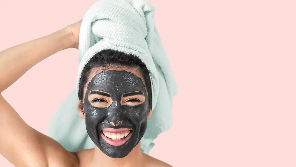 Happy smiling girl applying facial black mask - Young woman having skin care cleanser spa day - Beauty clean treatment and cosmetology products concept - Pink background