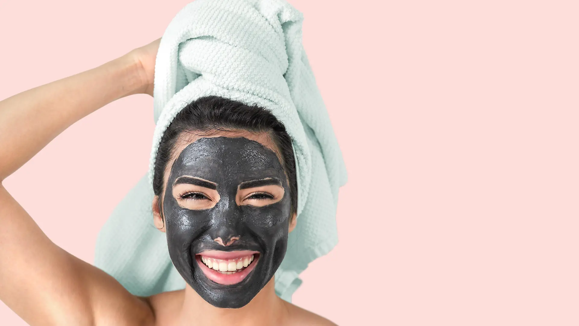 Happy smiling girl applying facial black mask - Young woman having skin care cleanser spa day - Beauty clean treatment and cosmetology products concept - Pink background