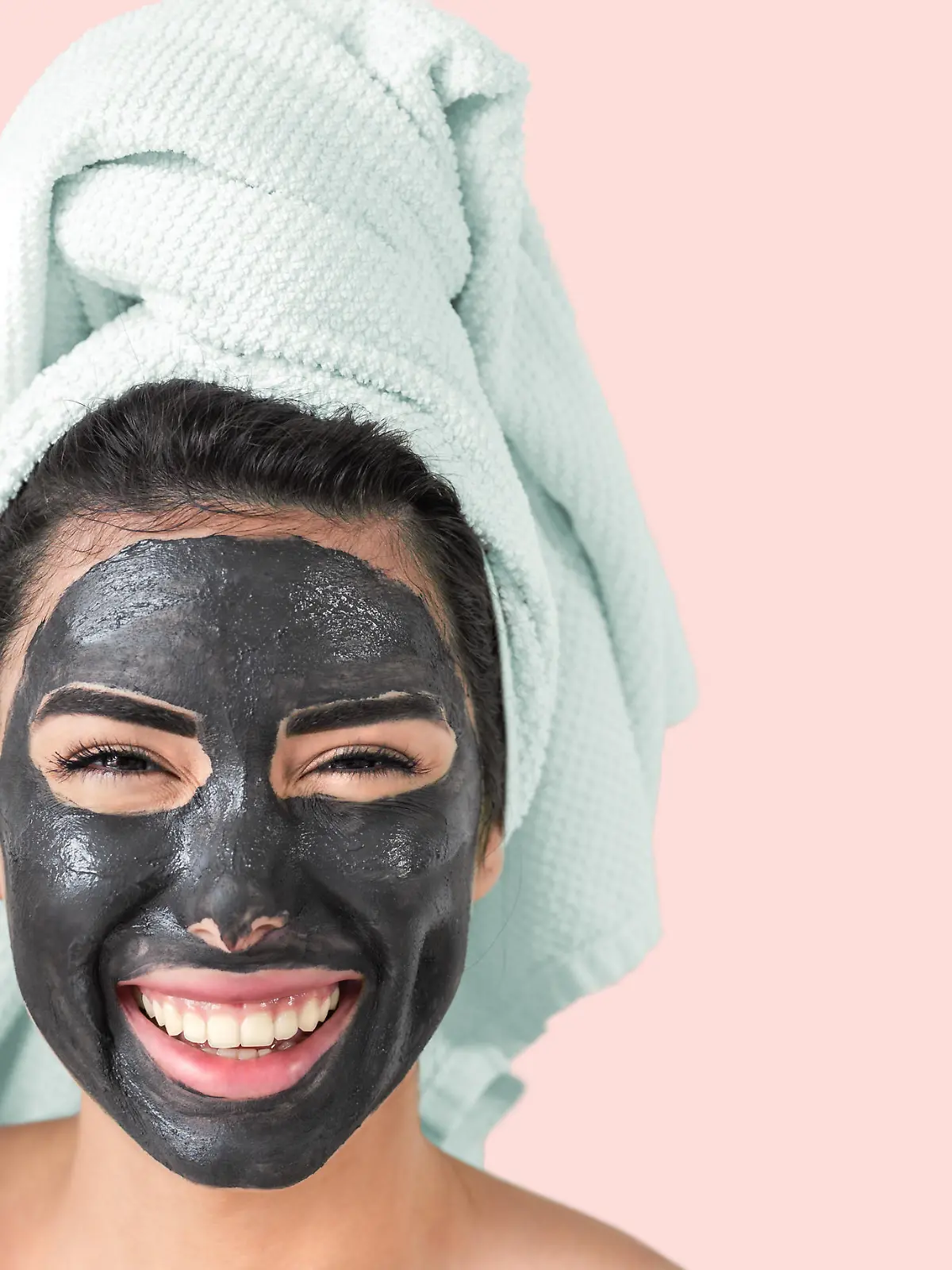 Happy smiling girl applying facial black mask - Young woman having skin care cleanser spa day - Beauty clean treatment and cosmetology products concept - Pink background