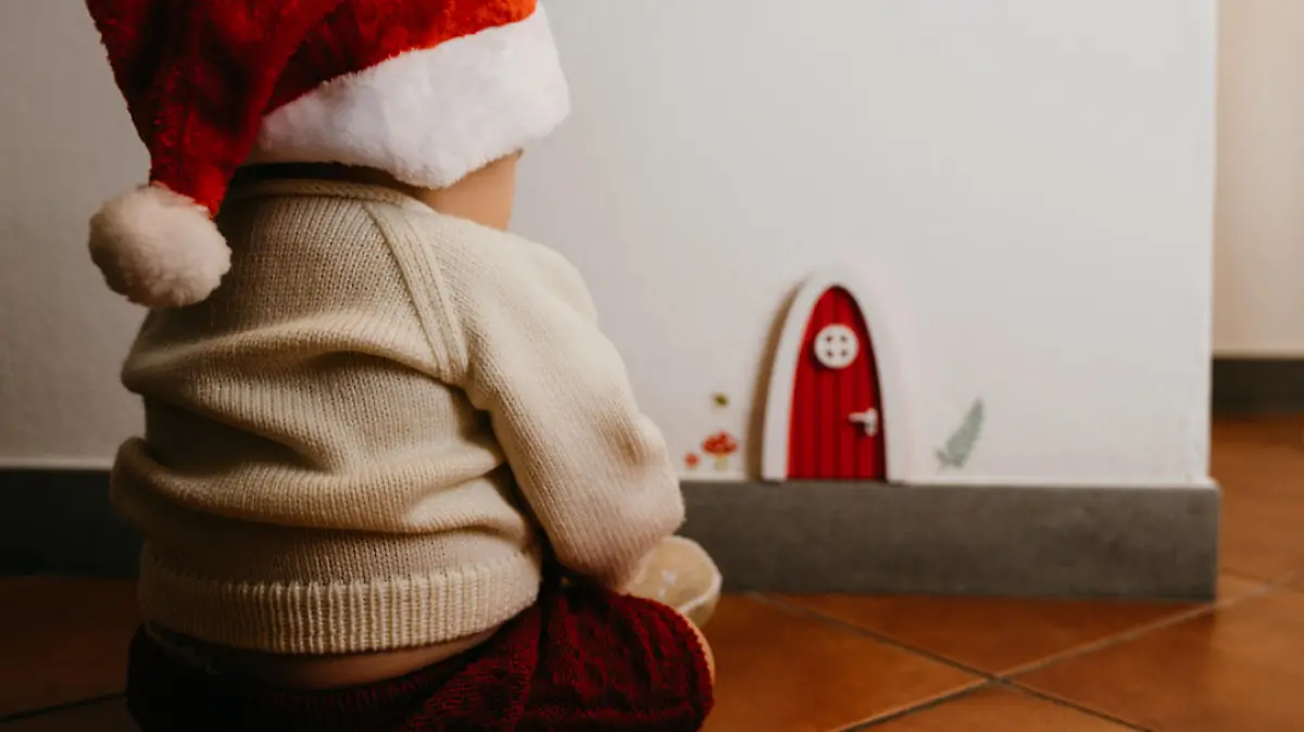 Little kid sitting near a gnomes door. He's wearing a santa hat.