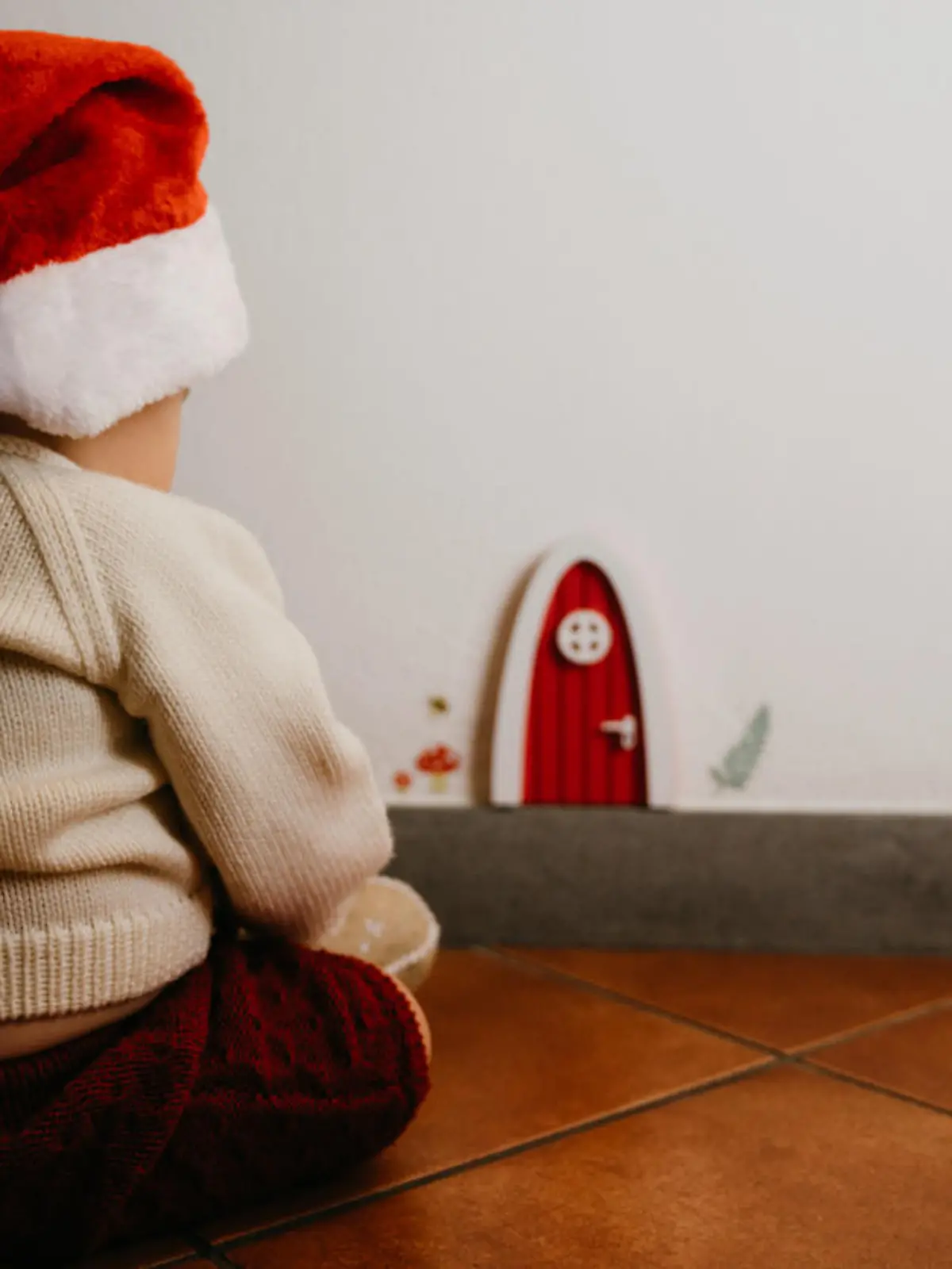 Little kid sitting near a gnomes door. He's wearing a santa hat.