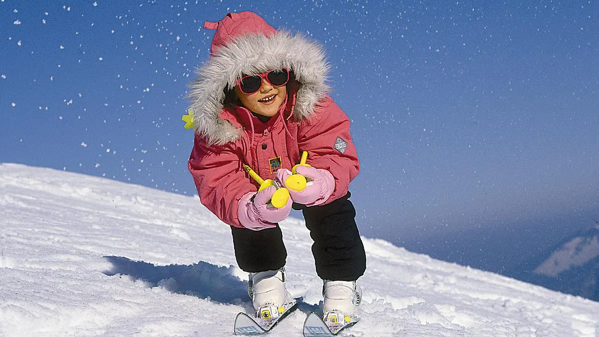 Ein Kind fährt in der Hocke auf seinen Abfahrtsskiern einen Schneeberg hinunter.