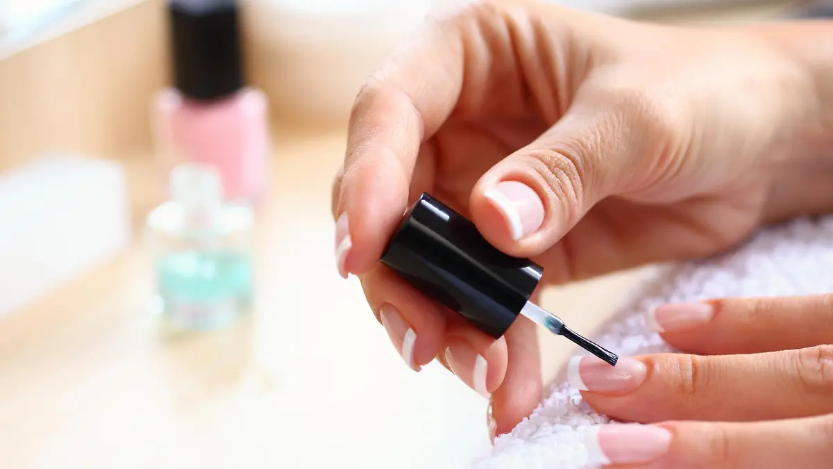Closeup of adult caucasian unrecognizable woman painting her fingernails with nail polish product. Her hands are placed on a white towel. blurry cosmetic products in background.