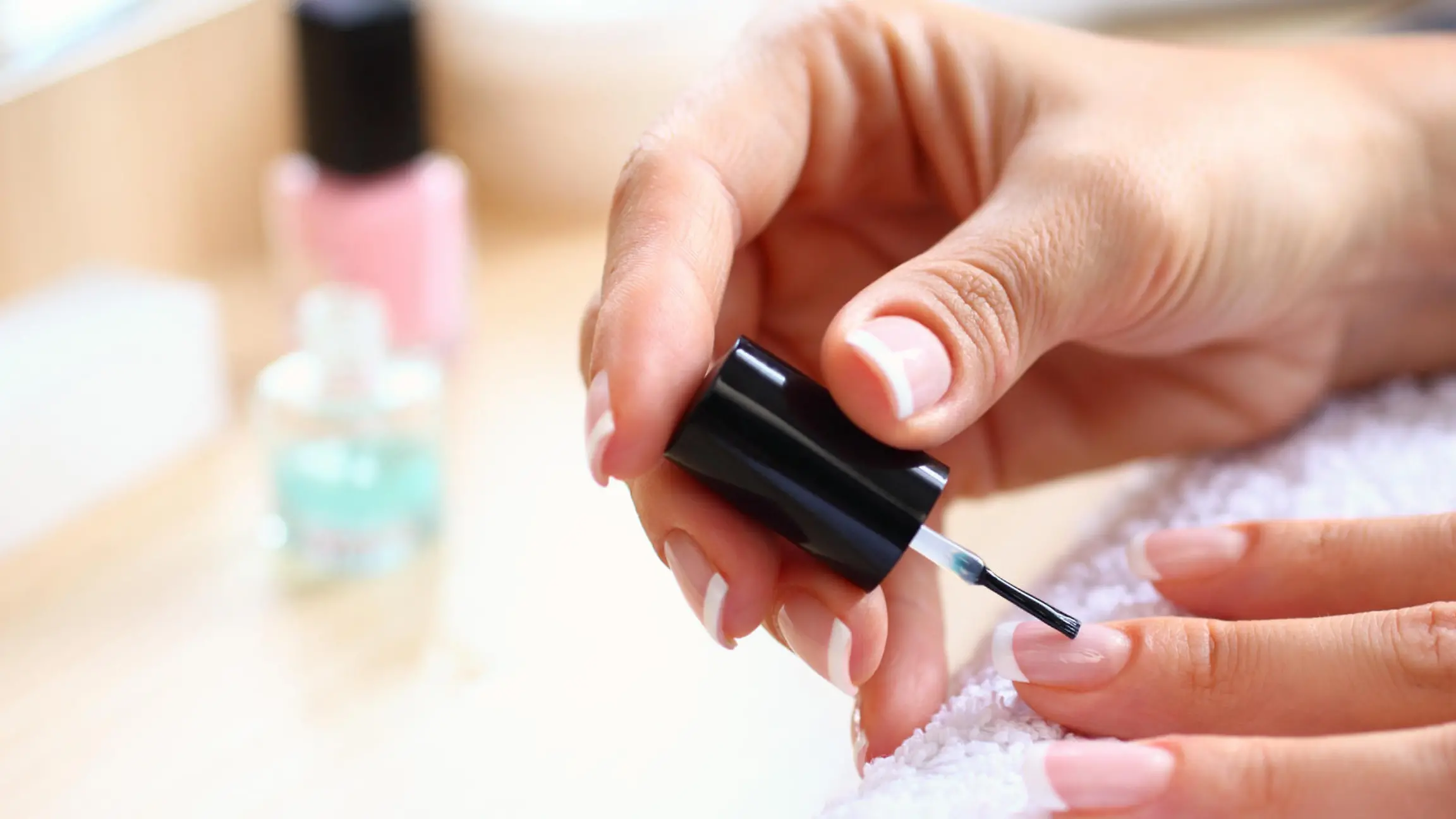Closeup of adult caucasian unrecognizable woman painting her fingernails with nail polish product. Her hands are placed on a white towel. blurry cosmetic products in background.