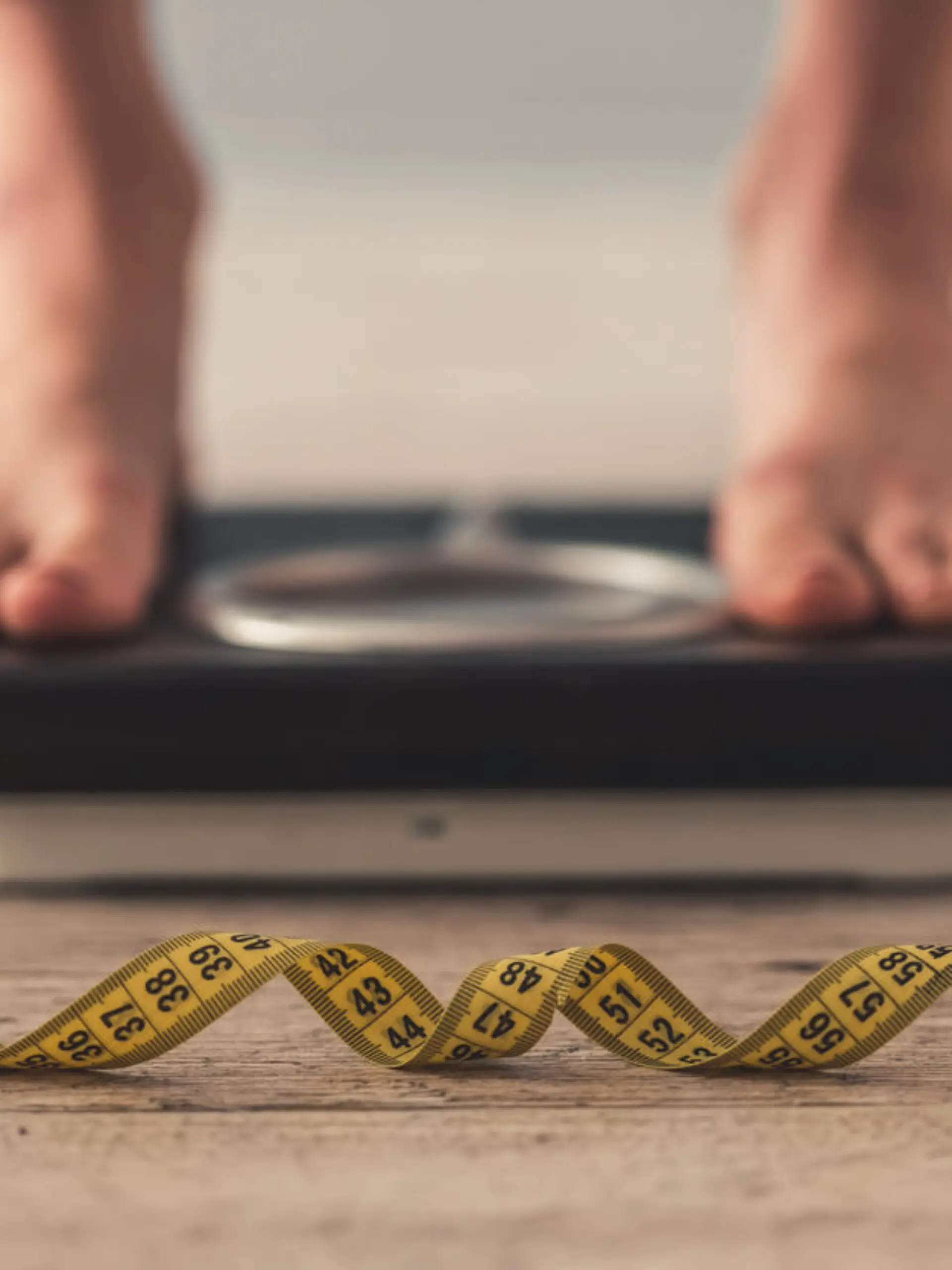 Cropped image of woman feet standing on weigh scales, on gray background. A tape measure in the foreground