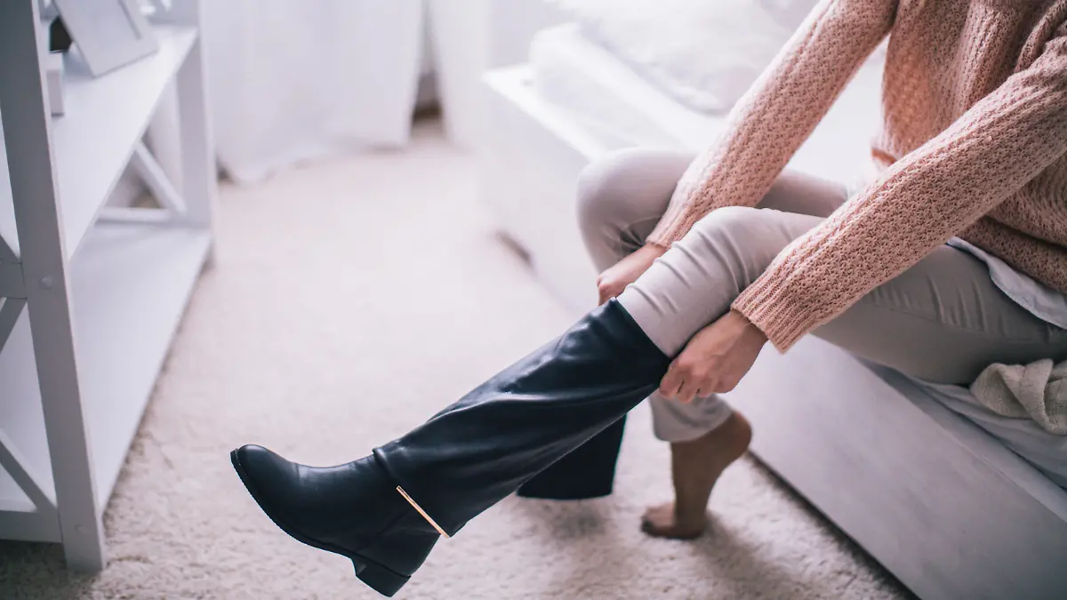 Young woman sitting at the bed in her bedroom, getting ready for work, putting on her boots.