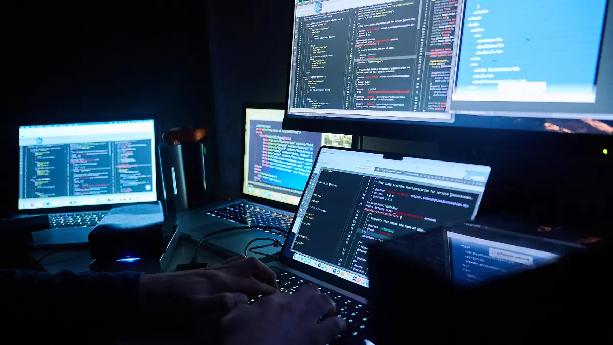 Illustration: Fiktive html-Seiten und Hacker-Programme sind auf Bildschirmen zu sehen, während ein Mann seinen Hände auf der Tastatur hat.