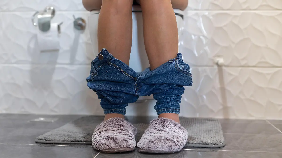 Frau sitzt in Jeans und Hausschuhen auf Toilette, nur der Unterkörper sichtbar.