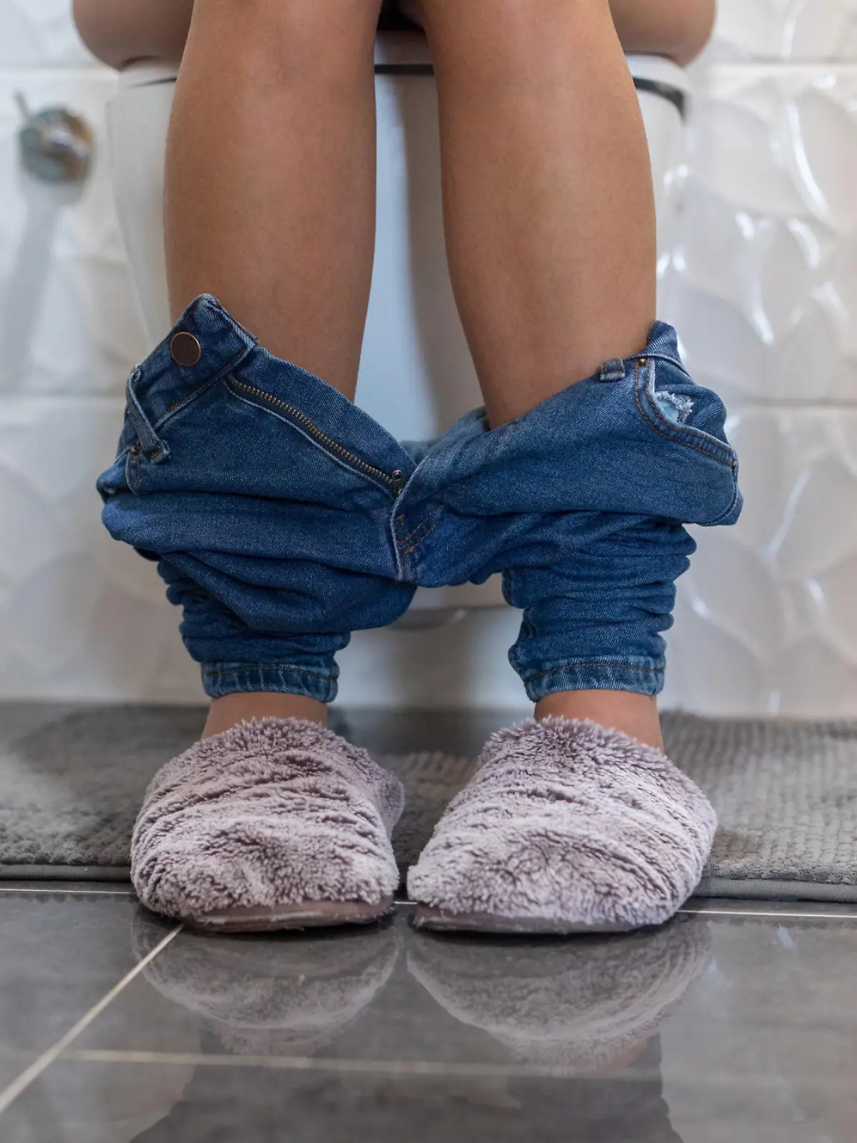 Frau sitzt in Jeans und Hausschuhen auf Toilette, nur der Unterkörper sichtbar.