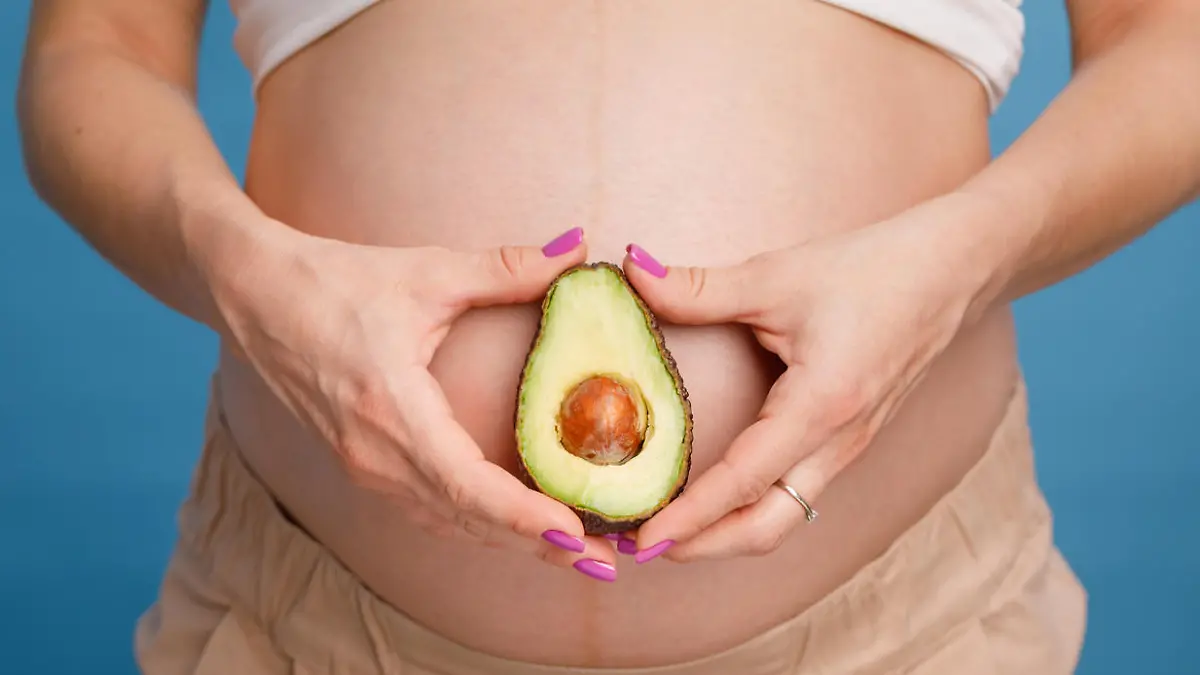 Schwangere Frau hält Avocado vor ihren Bauch.