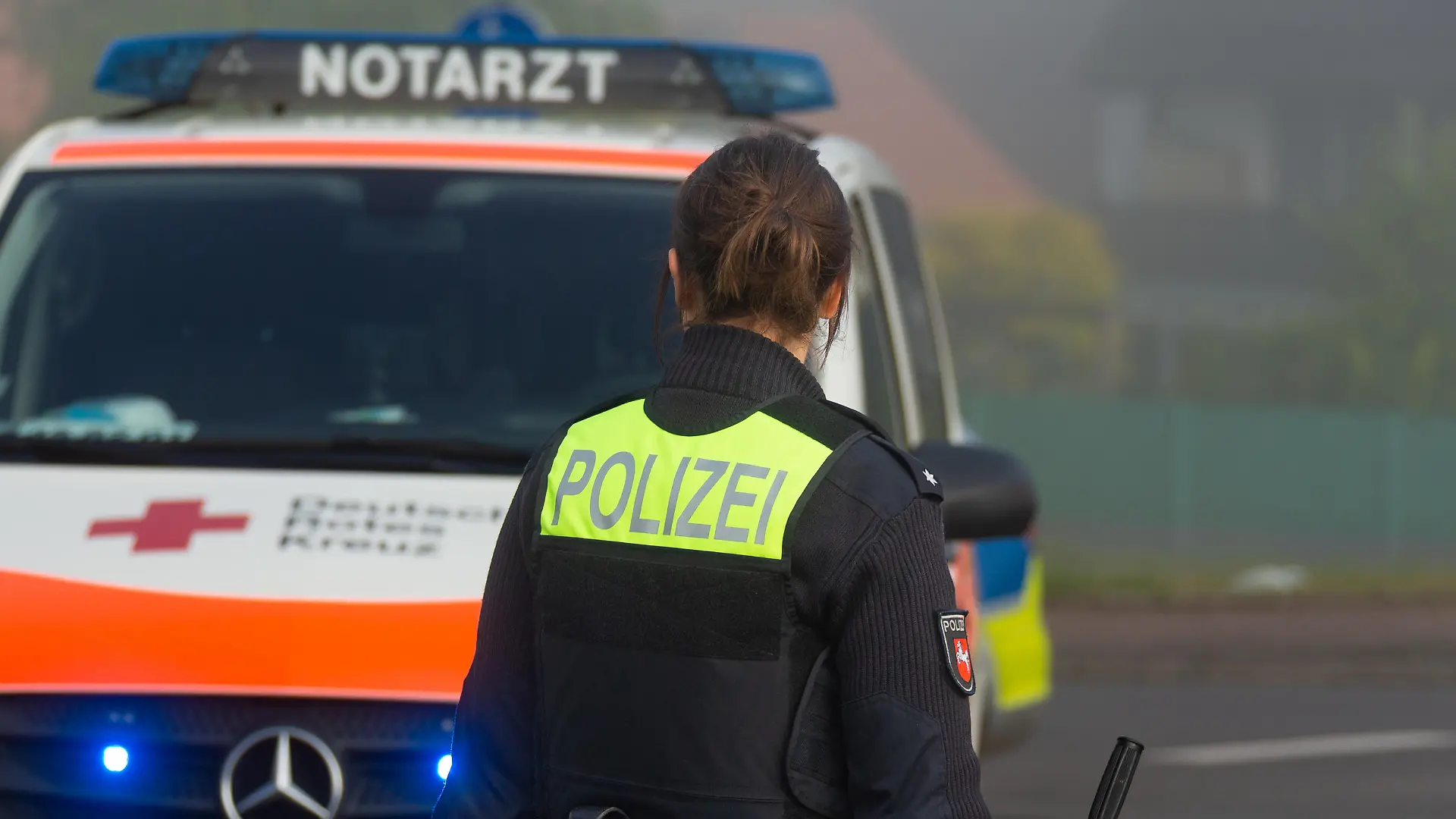 Melle, Deutschland 02. Juni 2022: Eine Beamtin der Polizei Niedersachsen steht an einem Unfallort, mit dem Schriftzug Polizei auf der Weste, im Hintergrund steht ein Einsatzfahrzeug vom DRK mit dem Schriftzug Notarzt und Blaulicht auf der Straße.