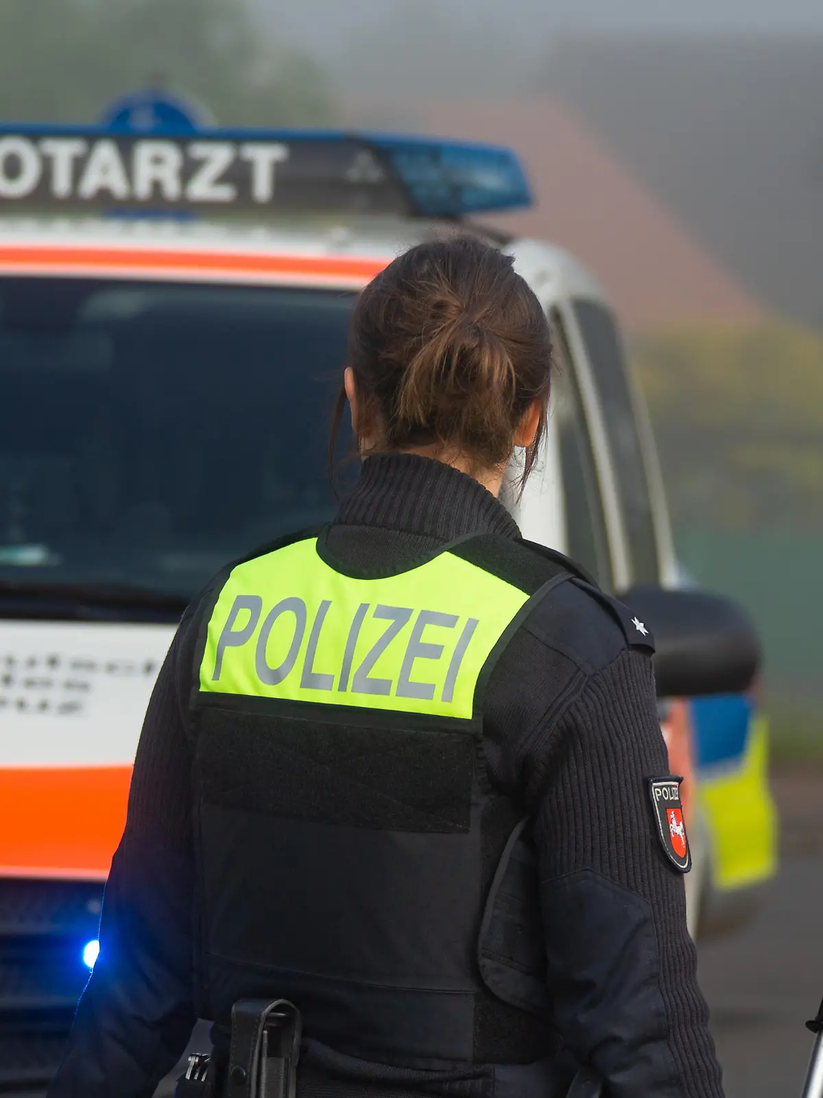 Melle, Deutschland 02. Juni 2022: Eine Beamtin der Polizei Niedersachsen steht an einem Unfallort, mit dem Schriftzug Polizei auf der Weste, im Hintergrund steht ein Einsatzfahrzeug vom DRK mit dem Schriftzug Notarzt und Blaulicht auf der Straße.