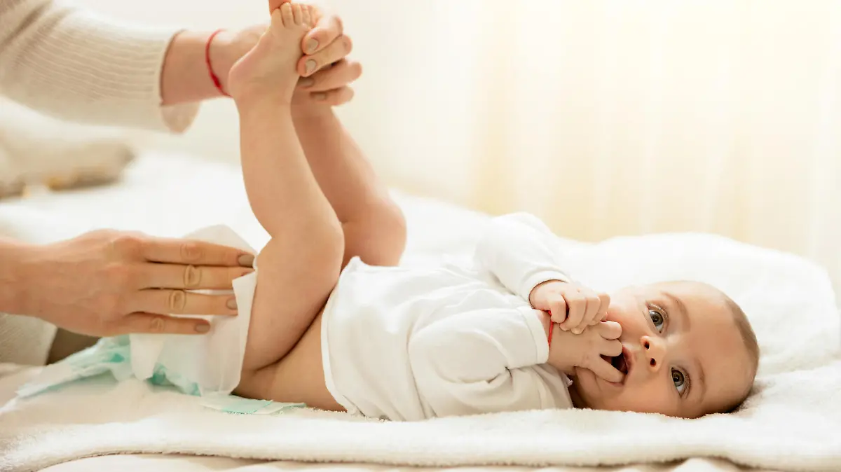 Baby boy lying on the bed with hands in the mouth while getting his diaper changed.
