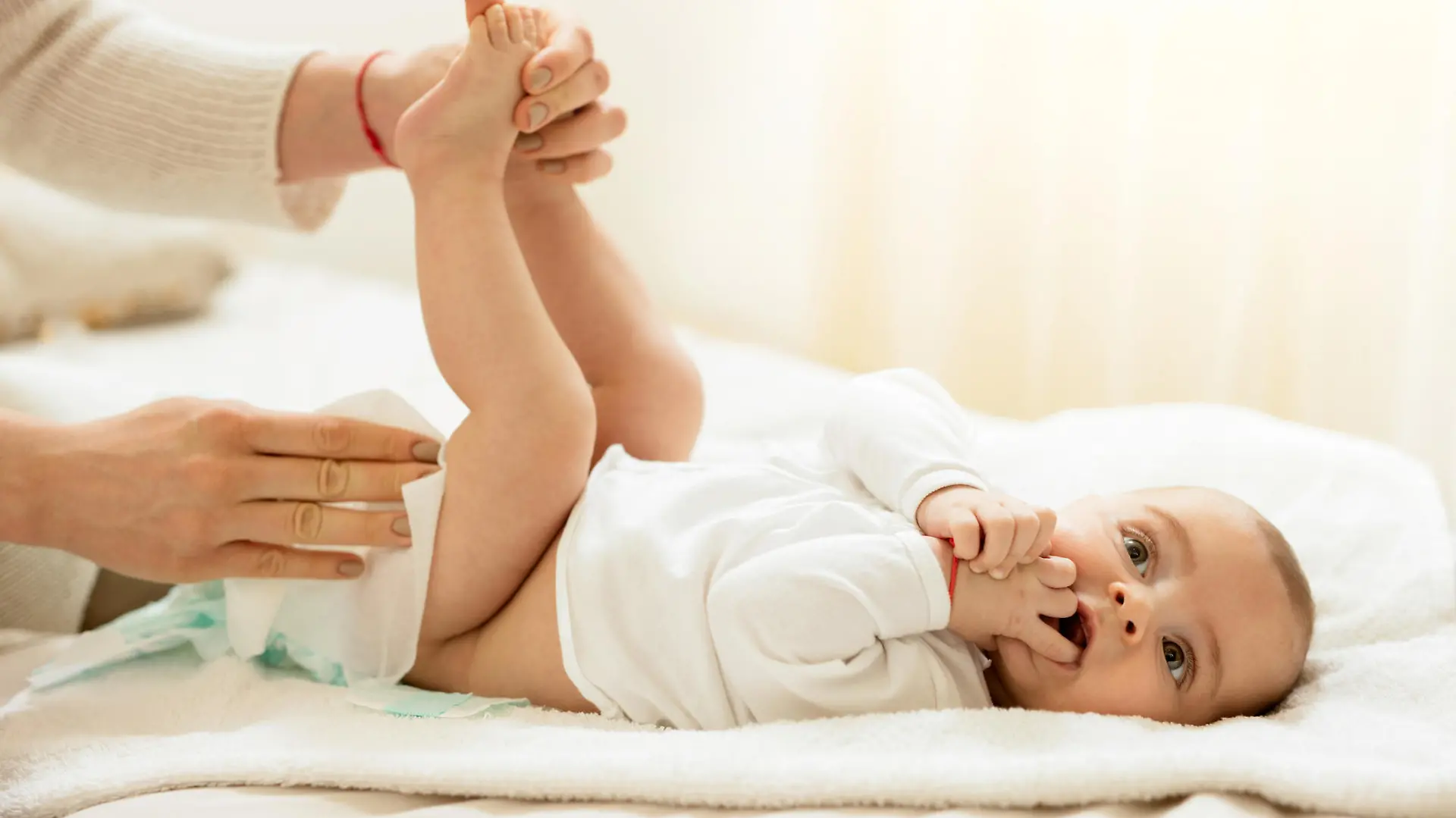 Baby boy lying on the bed with hands in the mouth while getting his diaper changed.