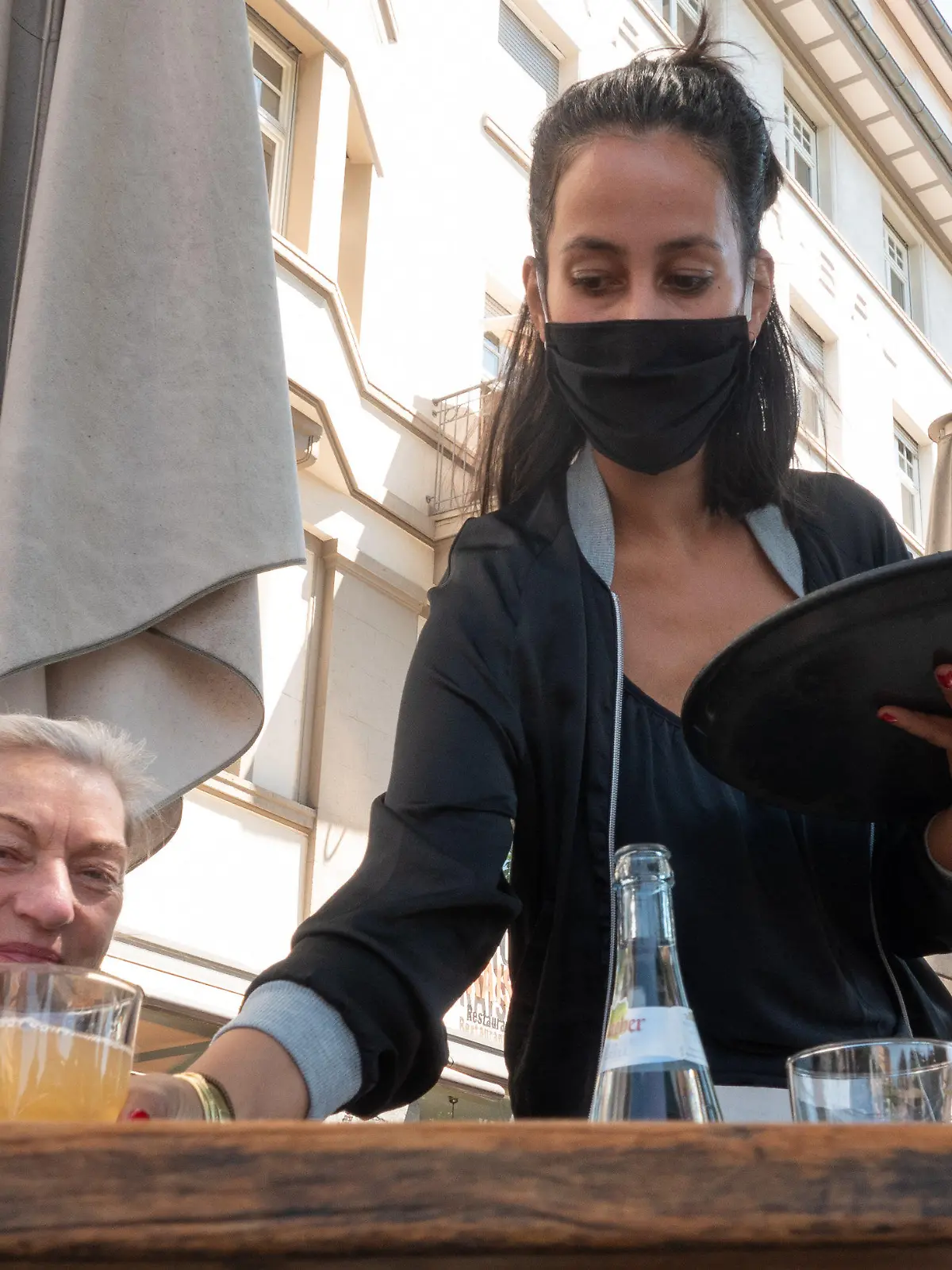 18.05.2020, Baden-Württemberg, Stuttgart: Eine Kellnerin mit Mundschutz stellt auf der Terrasse vor dem Café Kaiserbau einer Kundin ein Getränk auf den Tisch. Speise-Gaststätten, Ferienwohnungen und Campingplätze dürfen ab dem 18.05.2020 unter Einhaltung bestimmter Auflagen wieder für Ihre Gäste öffnen. Foto: Gregor Bauernfeind/dpa +++ dpa-Bildfunk +++