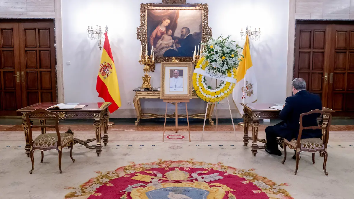 Ein Mann schreibt in der Apostolischen Nuntiatur in Madrid in ein offenes Kondolenzbuch neben einem Porträt des Papstes und einem Blumenkranz. 