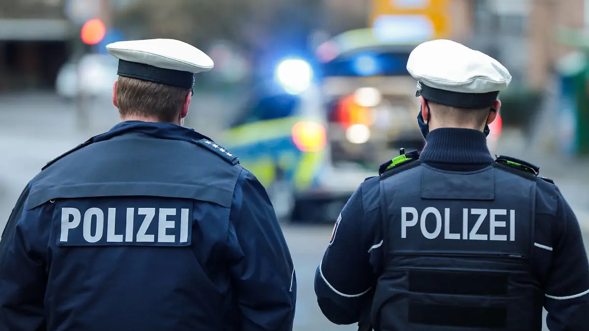 Symbolbild Einsatzgeschehen Ein Streifenwagen der Polizei NRW steht mit Blaulicht auf einer innerstädtischen Straße. Zwei Polizeibeamte mit Rückenaufschrift POLIZEI. *** Symbolic image of an operation A police patrol car of the North Rhine-Westphalia police force is parked on an inner-city street with blue lights flashing Two police officers with the inscription POLICE on their backs