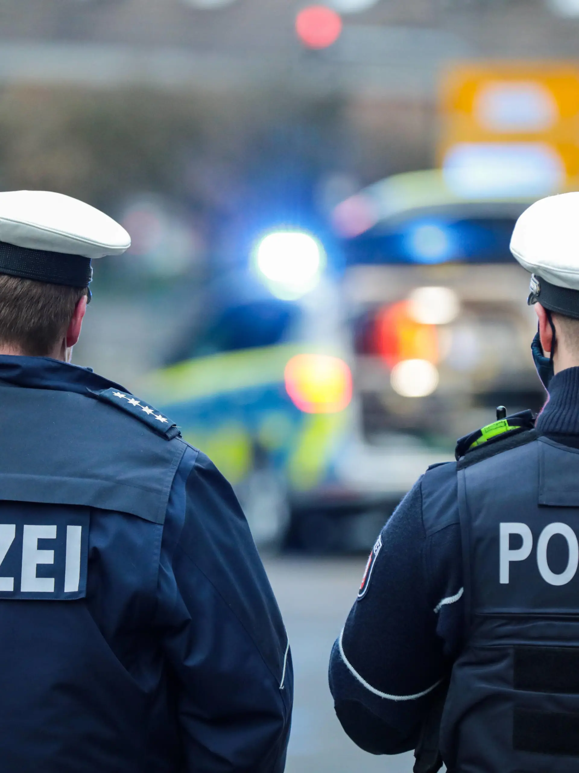 Symbolbild Einsatzgeschehen Ein Streifenwagen der Polizei NRW steht mit Blaulicht auf einer innerstädtischen Straße. Zwei Polizeibeamte mit Rückenaufschrift POLIZEI. *** Symbolic image of an operation A police patrol car of the North Rhine-Westphalia police force is parked on an inner-city street with blue lights flashing Two police officers with the inscription POLICE on their backs 