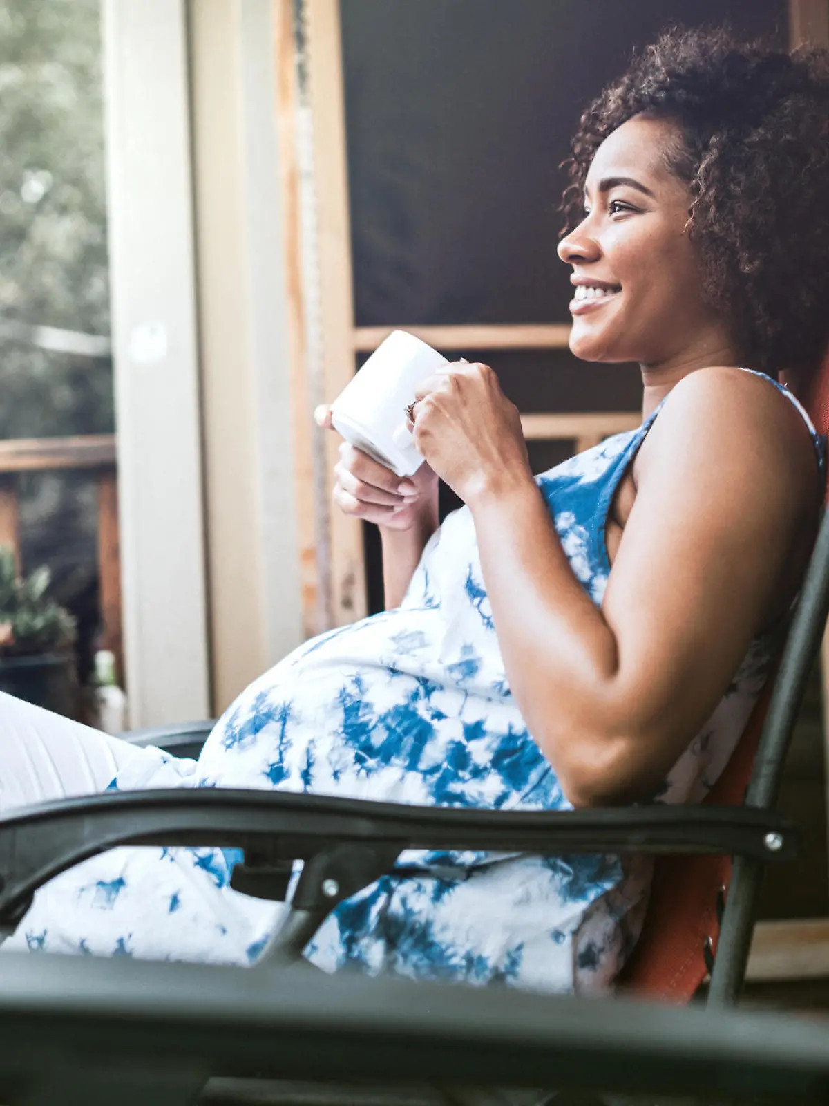 A beautiful African American woman in the 3rd trimester of her pregnancy rests in a recliner on her porch, enjoying a hot drink while anticipating her upcoming child birth.  Horizontal image with copy space.