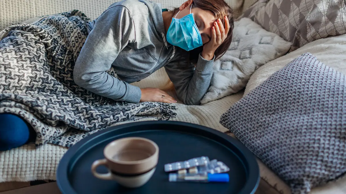 Sick woman having flu or cold. Girl lying in bed having headache wearing protective mask by pills and water on table.