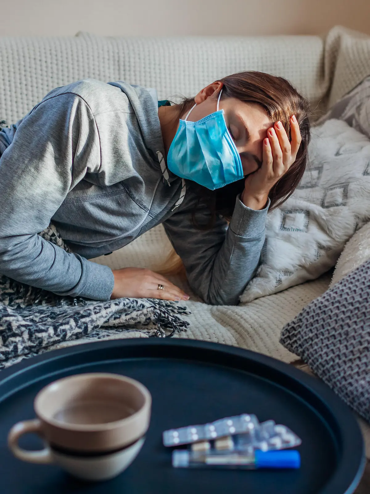 Sick woman having flu or cold. Girl lying in bed having headache wearing protective mask by pills and water on table.