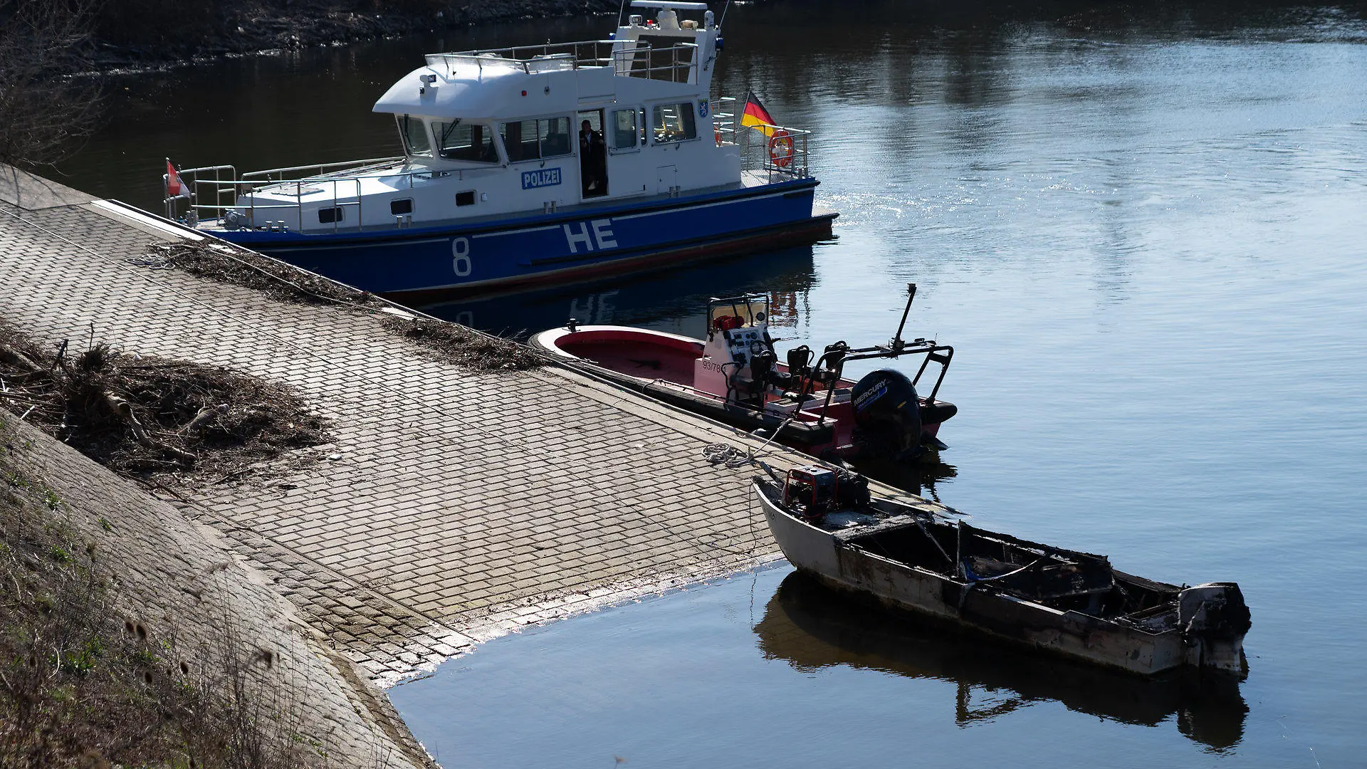 06.03.2022, Hessen, Wiesbaden: Ein ausgebranntes Boot (vorne) liegt vor einem Polizeiboot. Beim Brand eines Motorbootes am Sonntagmorgen im Rheinhafen von Wiesbaden-Schierstein ist ein siebenjähriges Mädchen ums Leben gekommen. Foto: Marvin Filipovic/5vision.media/dpa +++ dpa-Bildfunk +++