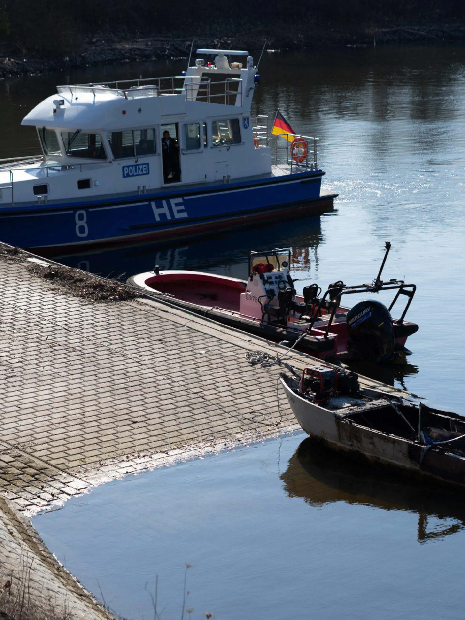 06.03.2022, Hessen, Wiesbaden: Ein ausgebranntes Boot (vorne) liegt vor einem Polizeiboot. Beim Brand eines Motorbootes am Sonntagmorgen im Rheinhafen von Wiesbaden-Schierstein ist ein siebenjähriges Mädchen ums Leben gekommen. Foto: Marvin Filipovic/5vision.media/dpa +++ dpa-Bildfunk +++