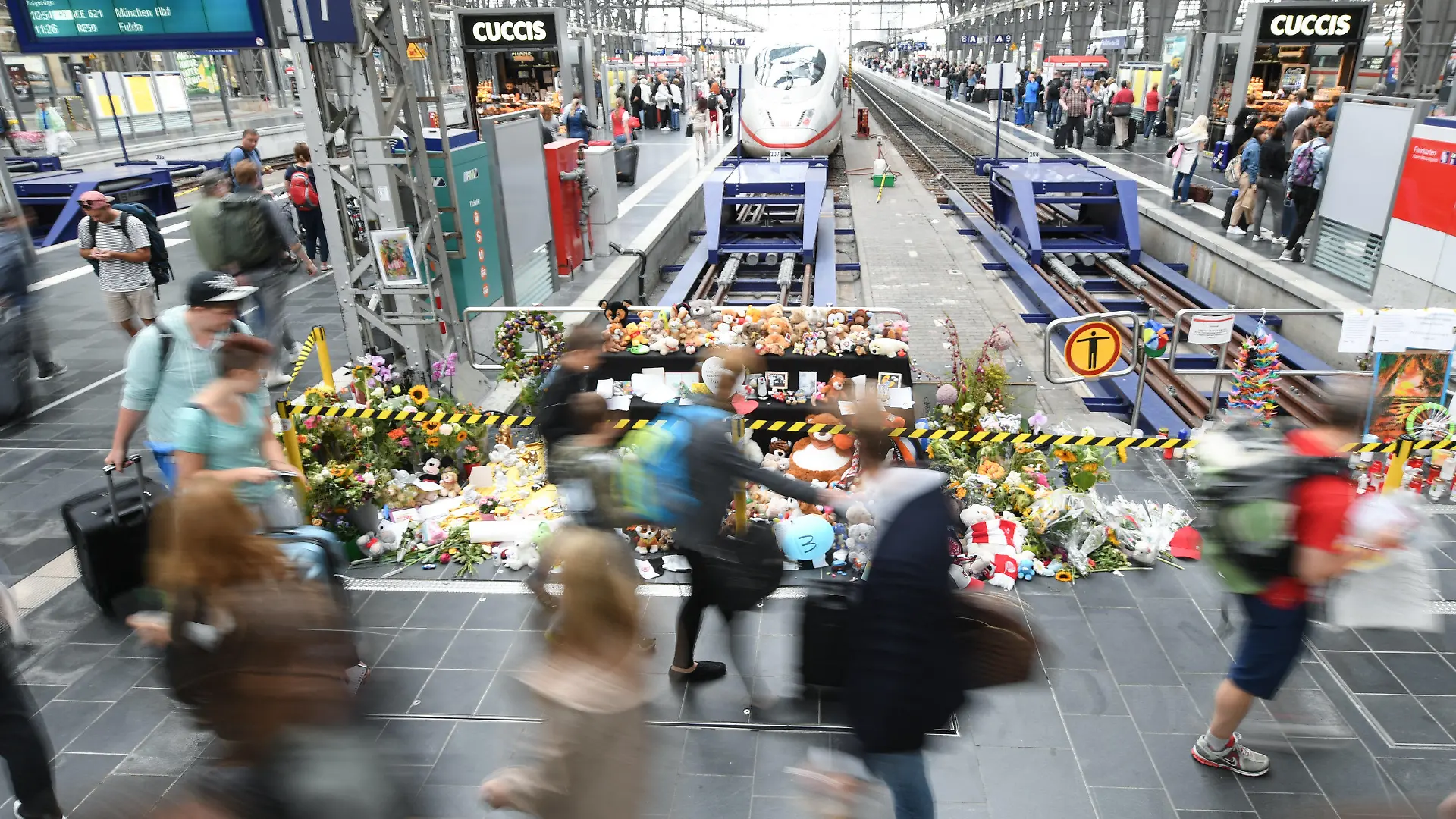 ARCHIV - 20.08.2019, Hessen, Frankfurt/Main: Passanten gehen am Gleis 7 des Hauptbahnhofs an einer Gedenkstelle vorbei, an der sich Blumen, Kuscheltiere und Beileidsbekundungen angesammelt haben (Aufnahme mit längerer Verschlusszeit). Am 29. Juli 2019 wurden ein acht Jahre alter Junge und seine Mutter in das Gleisbett gestoßen. Die Frau konnte sich gerade noch zur Seite retten, ihr Sohn wurde von einem ICE erfasst und starb. (zu dpa «Voraussichtlich Urteilsverkündung um tödliche Gleisattacke») Foto: Arne Dedert/dpa +++ dpa-Bildfunk +++