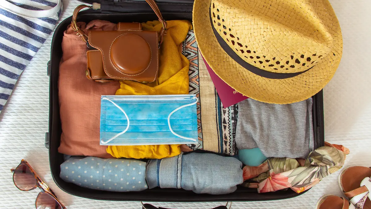 Suitcase prepared to travel in summer, in the new normal, after the coronavirus covid 19 pandemic, with a face mask among the luggage. There are other accessories like a hat, sunglasses, camera. Concept of quarantine, coronavirus, summer vacation in new normal. Flat lay or top view