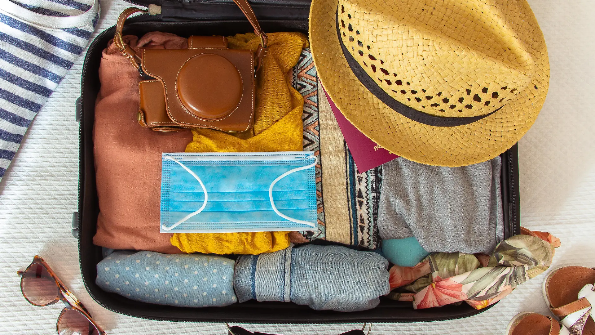 Suitcase prepared to travel in summer, in the new normal, after the coronavirus covid 19 pandemic, with a face mask among the luggage. There are other accessories like a hat, sunglasses, camera. Concept of quarantine, coronavirus, summer vacation in new normal. Flat lay or top view