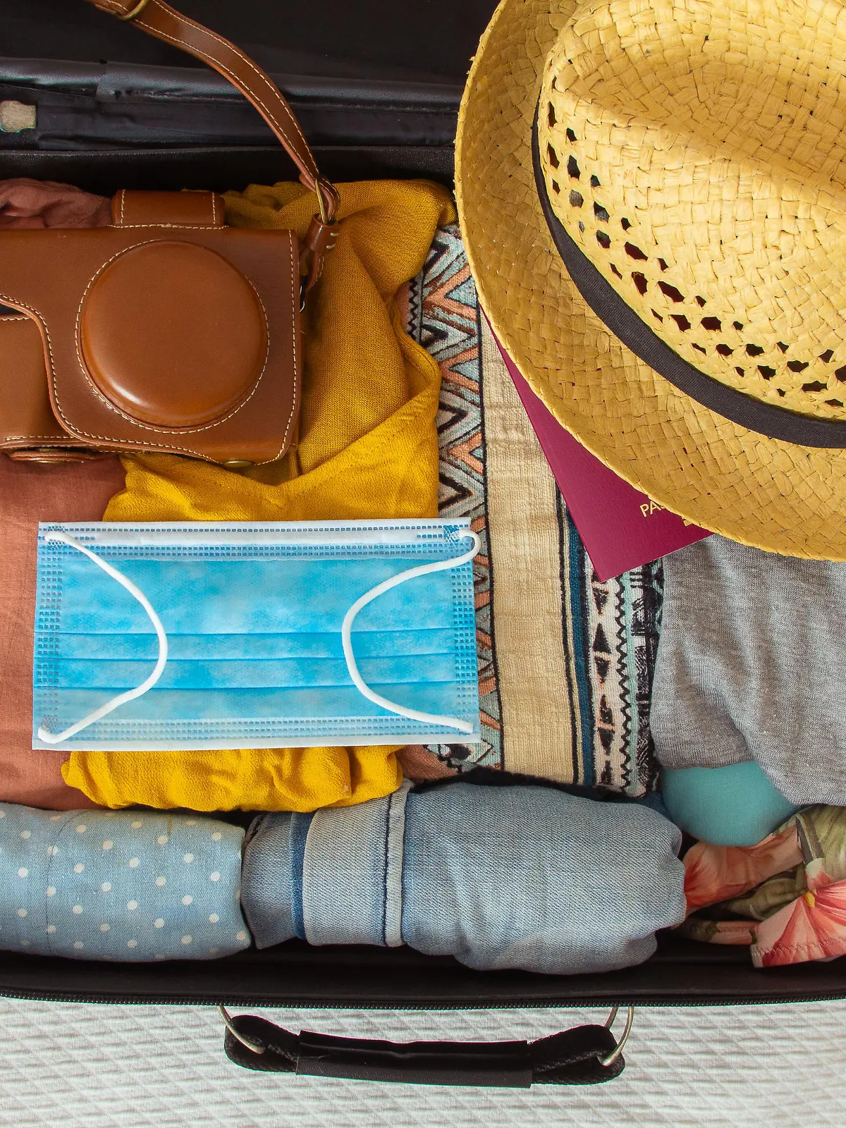 Suitcase prepared to travel in summer, in the new normal, after the coronavirus covid 19 pandemic, with a face mask among the luggage. There are other accessories like a hat, sunglasses, camera. Concept of quarantine, coronavirus, summer vacation in new normal. Flat lay or top view