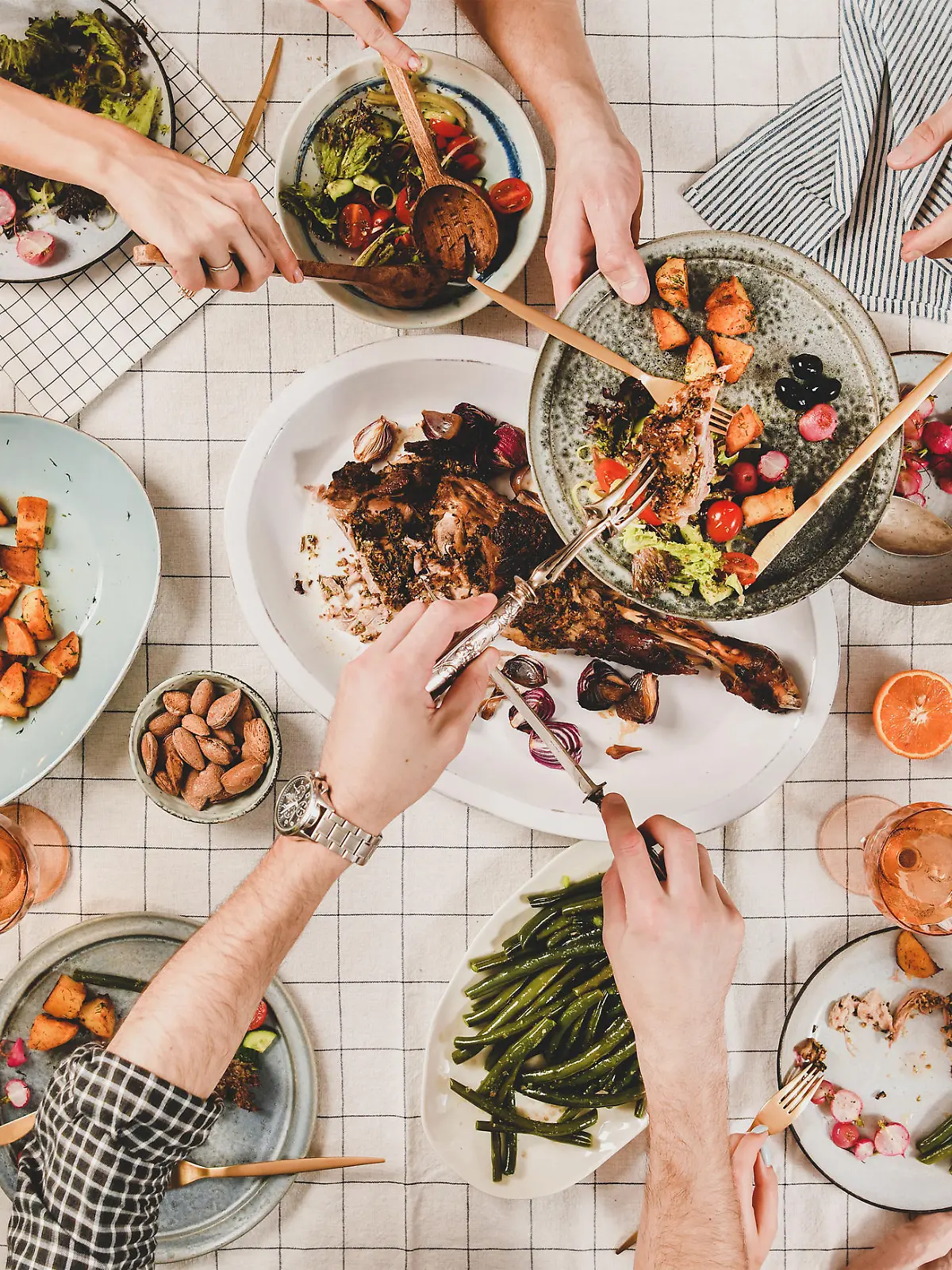 Family or friends gathering dinner. Flat-lay of peoples hands, roasted lamb shoulder, salads, vegetables, rose wine, mimosa branches over white checkered tablecloth, top view. Celebration party dinner
