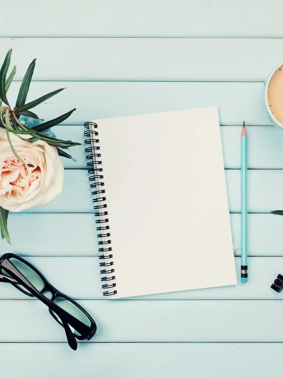 Morning coffee cup, clean notebook, pencil, eyeglasses and vintage rose flower in vase on blue rustic table top view. Planning and design concept. Cozy breakfast. Flat lay styling.