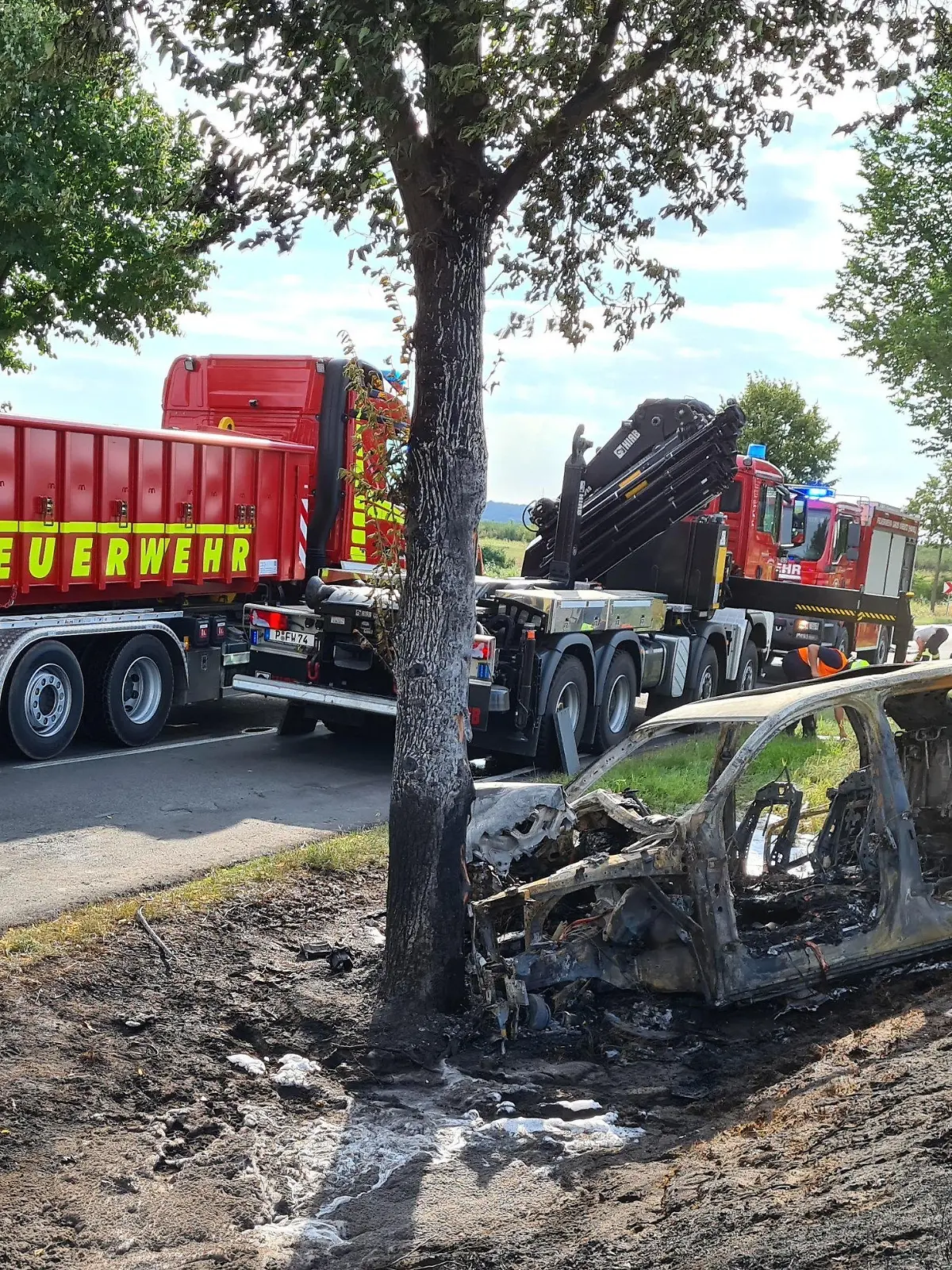 28.07.2020, Brandenburg, Groß Kreutz: Das ausgebrannte Unfallwrack eines Autos steht an einem Baum. Eine junge Fahrerin ist in Groß Kreutz bei Potsdam mit dem Auto gegen einen Baum geprallt und darin verbrannt. Es sei schwierig gewesen, die Frau in dem Elektroauto zu bergen, sagte ein Polizeisprecher. Feuerwehrleute hätten versucht, mit dem Kühlen von Akkus zu verhindern, dass die Batterien in dem Fahrzeug explodieren. (zu dpa «Fahrerin fährt gegen Baum und verbrennt im Auto») Foto: Julian Stähle/dpa-Zentralbild/dpa +++ dpa-Bildfunk +++