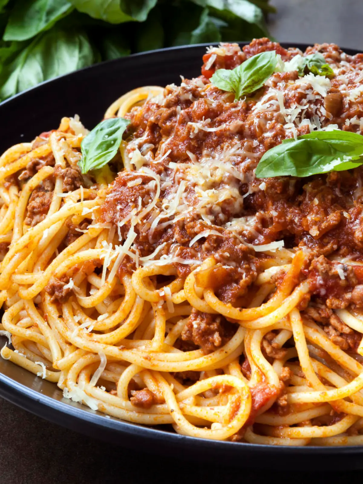 Spaghetti bolognese in black serving platter, with fresh basil and parmesan.
