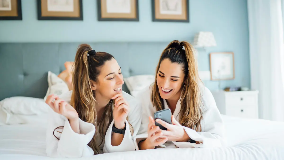  Smiling twin sisters using smart phone while lying on bed in hotel model released Symbolfoto property released OCMF01557