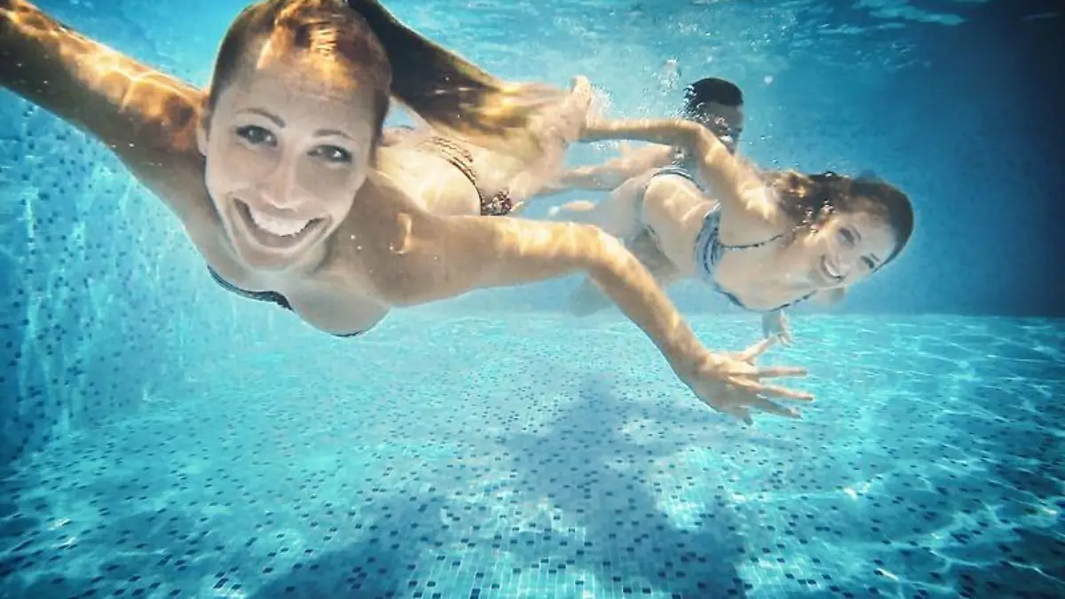 Group of mid 20's people taking underwater selfies in a swimming pool. There are two girls and a guy in foreground, keeping airtight, smiling and looking at camera.