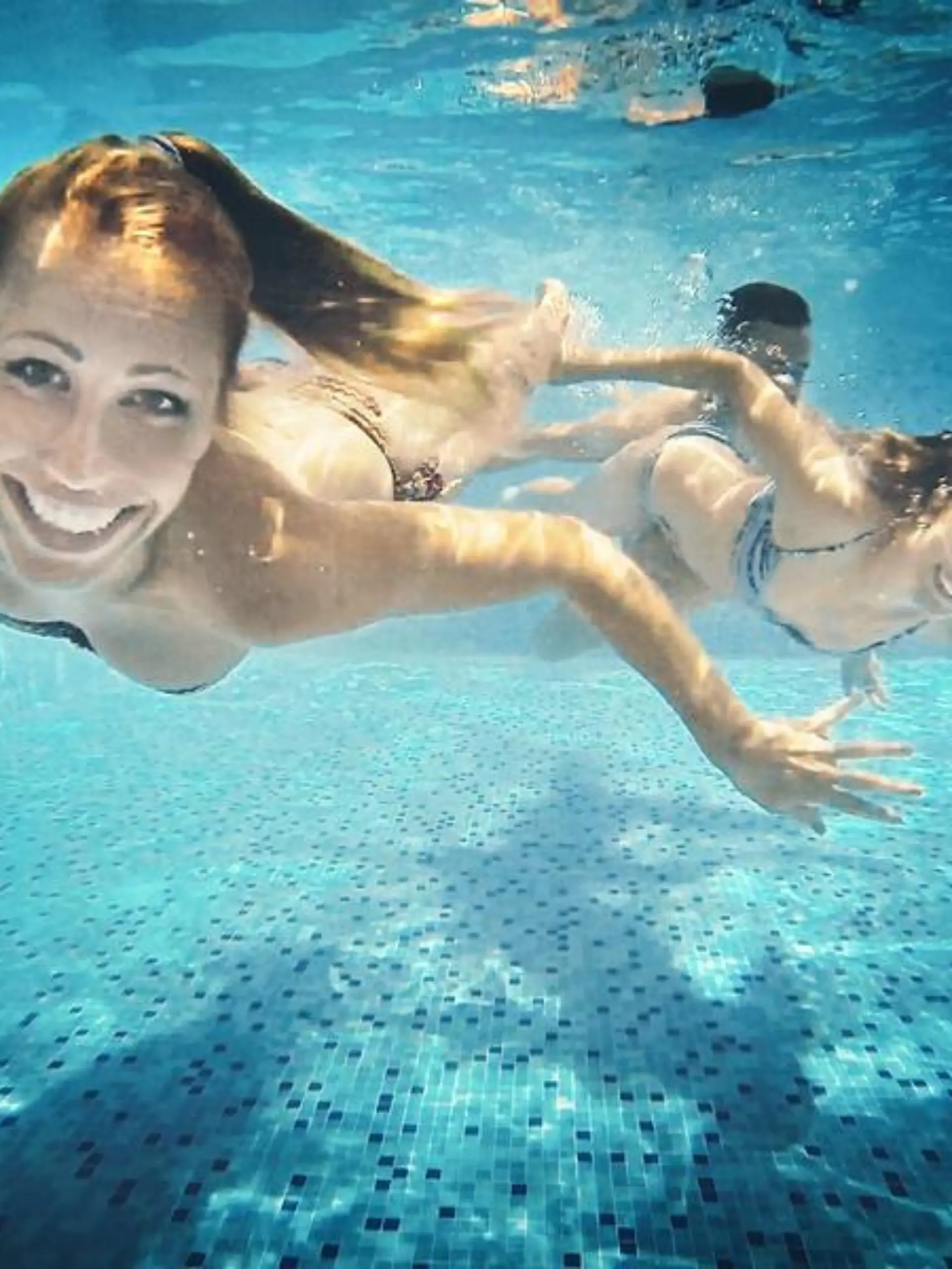 Group of mid 20's people taking underwater selfies in a swimming pool. There are two girls and a guy in foreground, keeping airtight, smiling and looking at camera.
