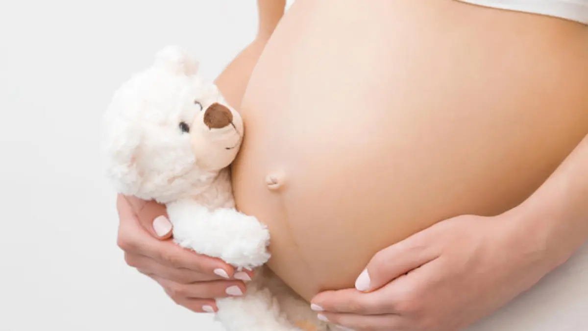schwangerer Teenager hält sich den Babybauch und hat einen Plüschteddy in der Hand
