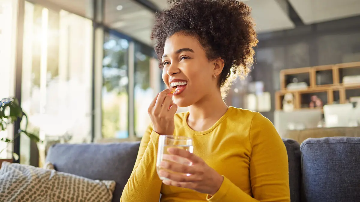 Young happy mixed race woman taking medication with water at home. One hispanic female with a curly afro taking a vitamin for good health while sitting on the couch at home. Woman drinking a supplement