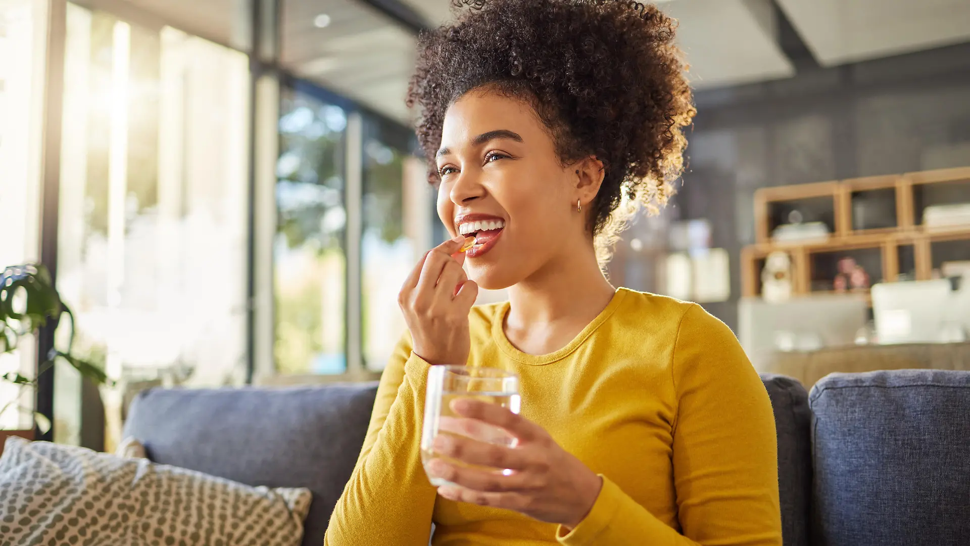 Young happy mixed race woman taking medication with water at home. One hispanic female with a curly afro taking a vitamin for good health while sitting on the couch at home. Woman drinking a supplement