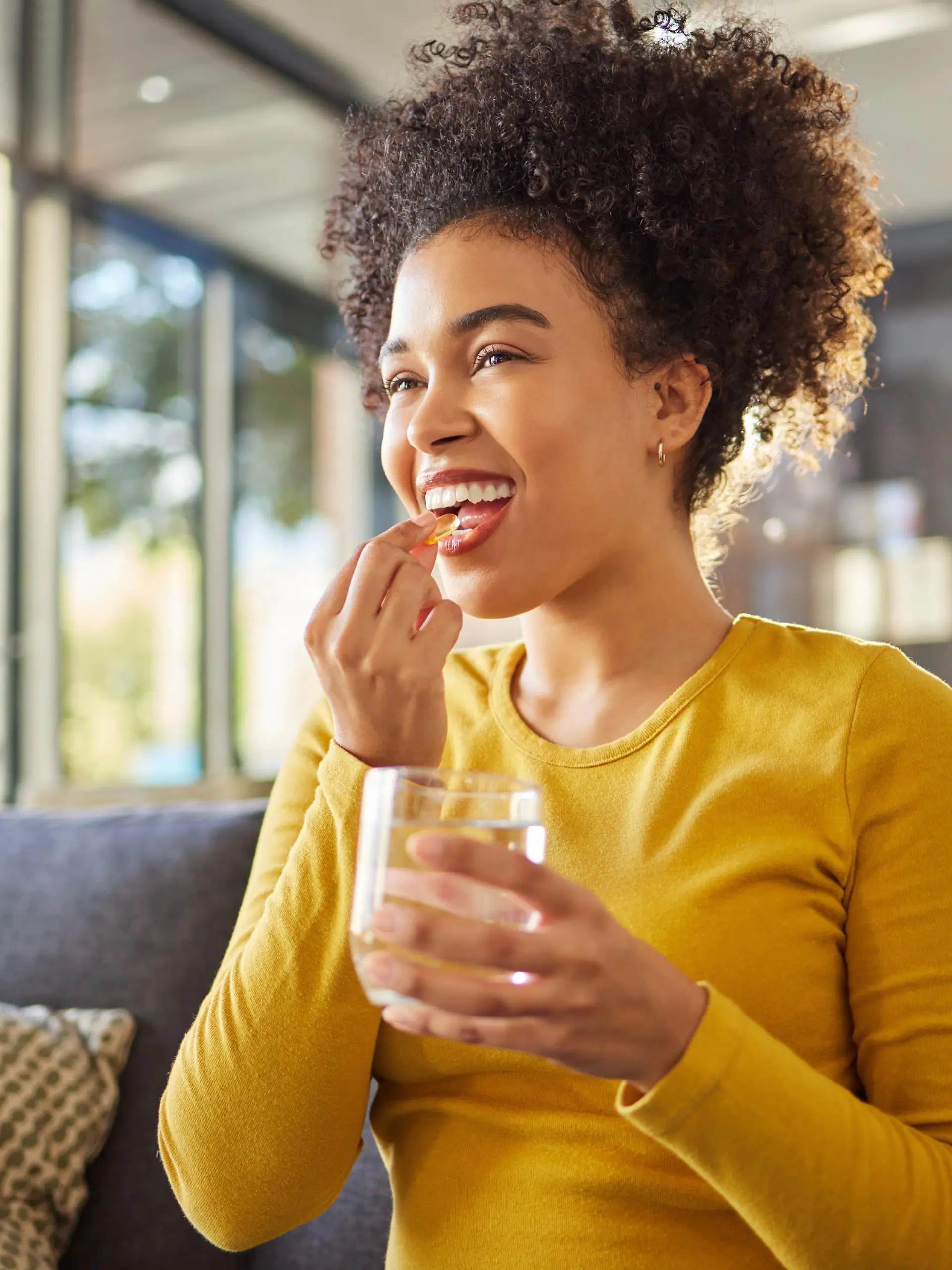 Young happy mixed race woman taking medication with water at home. One hispanic female with a curly afro taking a vitamin for good health while sitting on the couch at home. Woman drinking a supplement